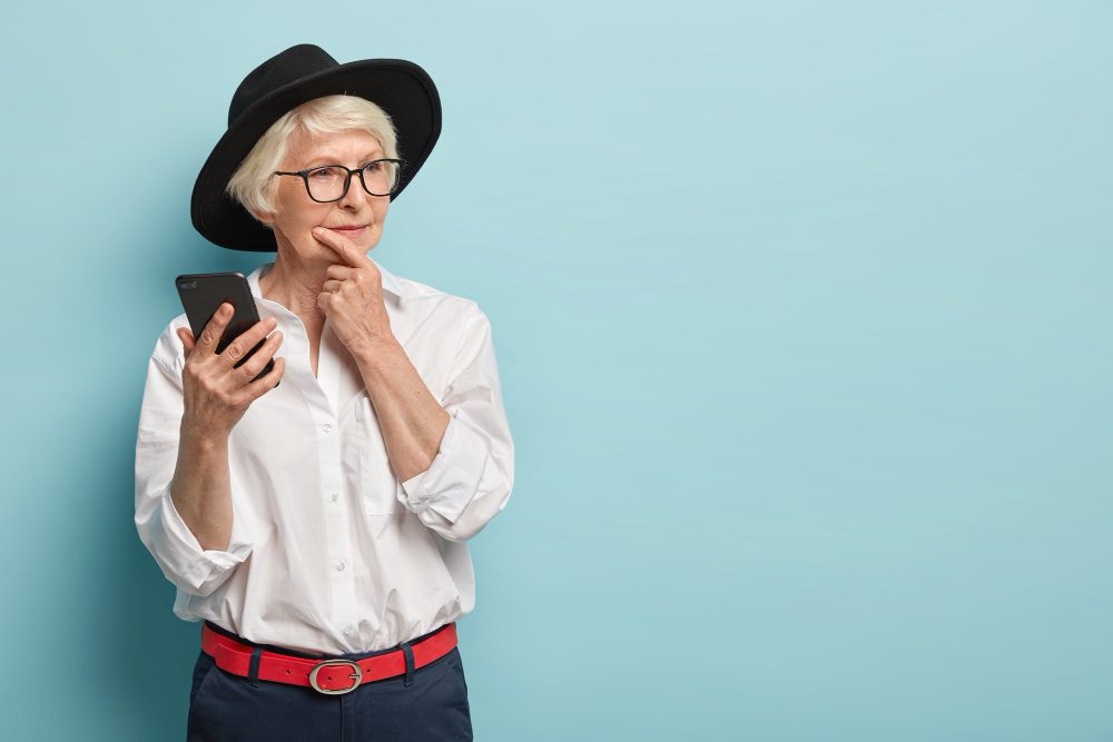 A stylish senior woman with white hair, wearing a black hat and glasses, holding a smartphone and looking thoughtfully to the side against a light blue background.